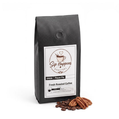A black bag of Sip Happens Roasters Pecan Pie Fresh Roasted Coffee stands on a white background with roasted coffee beans and pecans placed in front of the bag.
