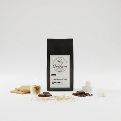 A black bag of Sip Happens Coffee Fresh Roasted Coffee stands centered on a white studio background, surrounded by s'mores-themed props including graham crackers, a small candle, chocolate pieces, cocoa powder, and marshmallows.