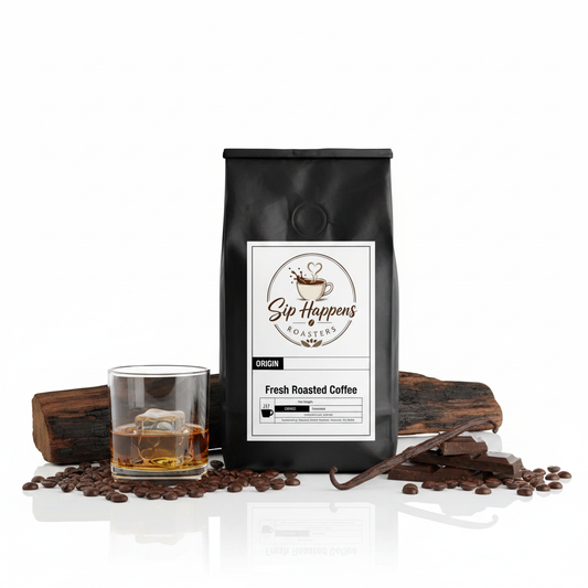 A black bag of Sip Happens Roasters Whiskey Barrel aged coffee stands on a white reflective surface, styled with coffee beans, a glass of amber coffee, vanilla bean, and chocolate pieces.